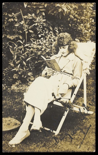 Download the full-sized image of A sailor in drag, wearing a long coat and hat, sits reading in a garden. Photographic postcard, ca. 1920.