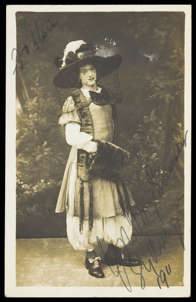 Download the full-sized image of A man in drag wearing a very large hat with a veil, stands in front of a painted backdrop, with dense foliage. Photographic postcard, 1911.