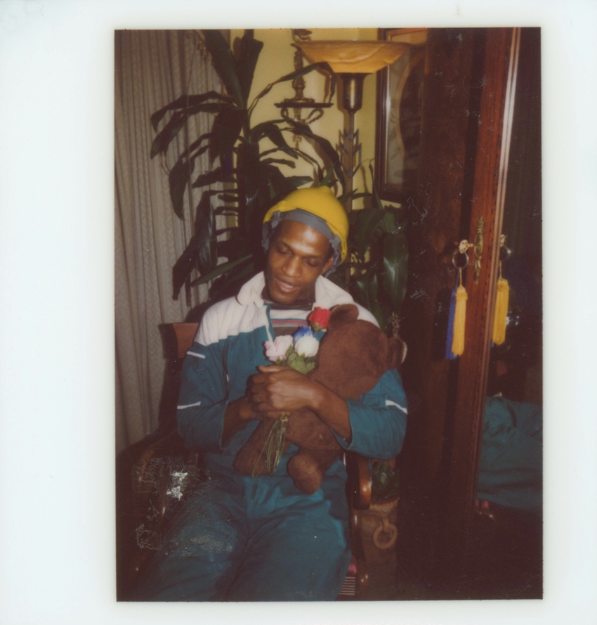 Download the full-sized image of A Photograph of Marsha P. Johnson Wearing a Construction Hat and Tracksuit, Holding a Stuffed Bear and Flowers