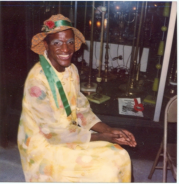 Download the full-sized image of A Photograph of Marsha P. Johnson Wearing a Yellow Floral Dress and a Patterned Orange Hat