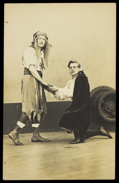 Download the full-sized image of Two amateur actors embrace on the deck of a ship; one is in drag, the other is kneeling on one knee. Photographic postcard, 191-.