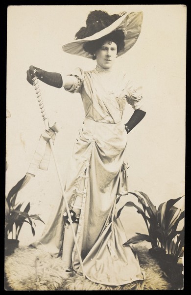 Download the full-sized image of An man in drag is wearing a revealing dress and holding a staff, posing with his other hand on his hip. Photographic postcard, 1909.