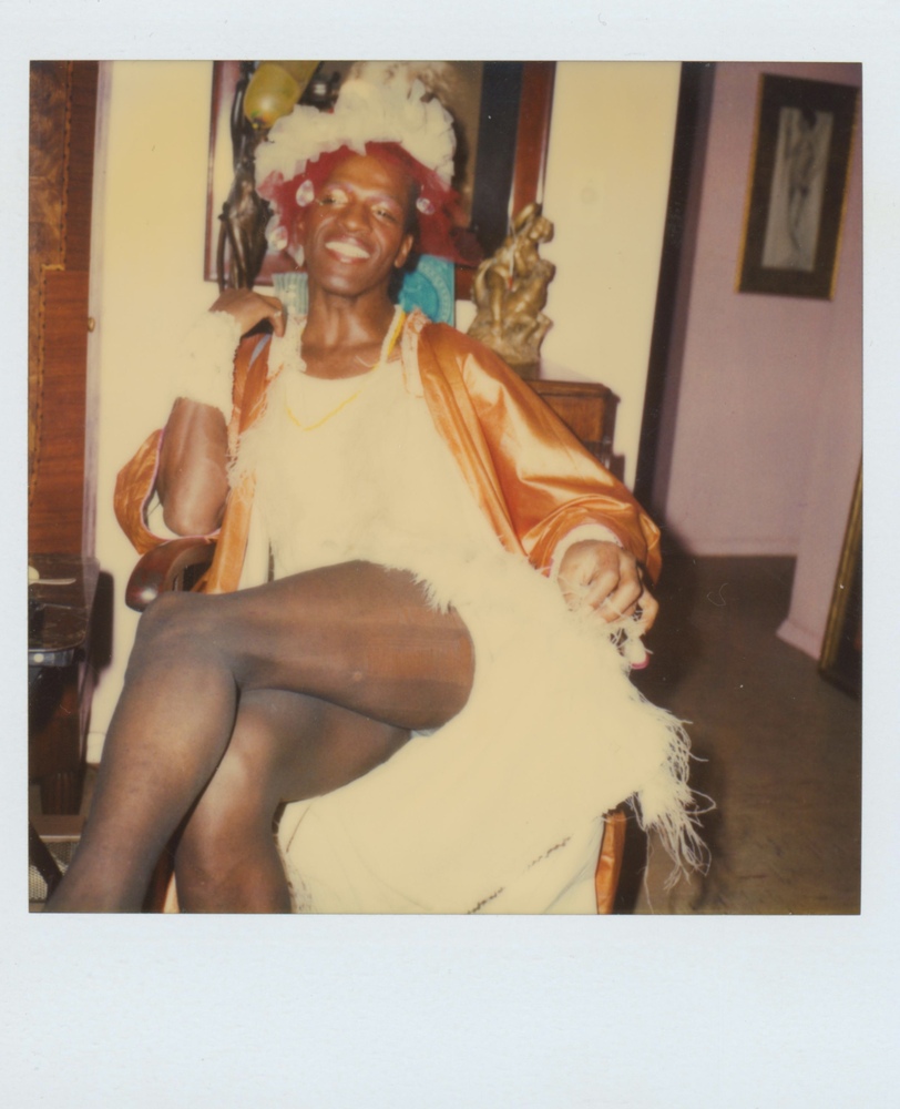 Download the full-sized image of A Photograph of Marsha P. Johnson Sitting With Her Legs Crossed, Smiling Wearing a Tulle Headpiece and in a White Orange Dress