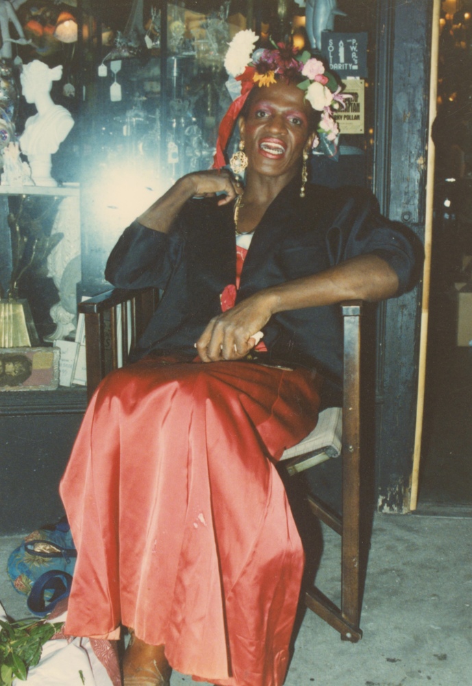 Download the full-sized image of A Photograph of Marsha P. Johnson Sitting Wearing a Floral Headpiece, Blue Jacket, and Red Dress