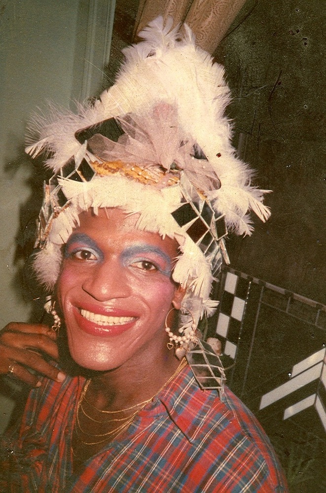 Download the full-sized image of A Photograph of Marsha P. Johnson Wearing a Feather Headpiece, Blue Eye Makeup, and Plaid Shirt