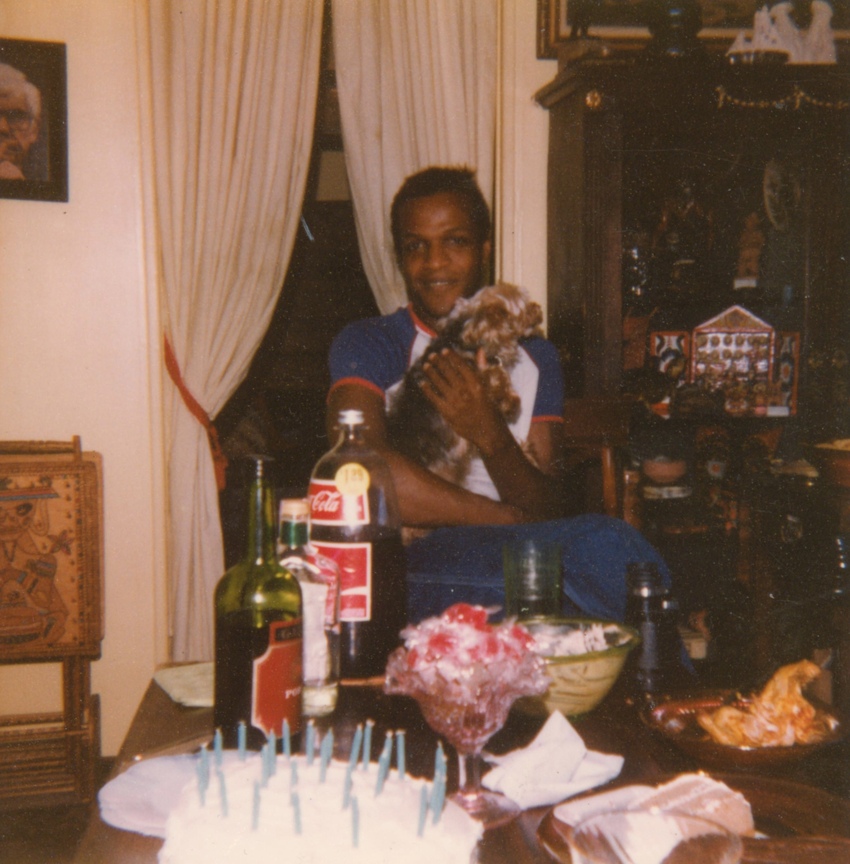 Download the full-sized image of A Photograph of Marsha P. Johnson Holding a Dog in Her Lap, Sitting at a Table with Party Food and Drinks