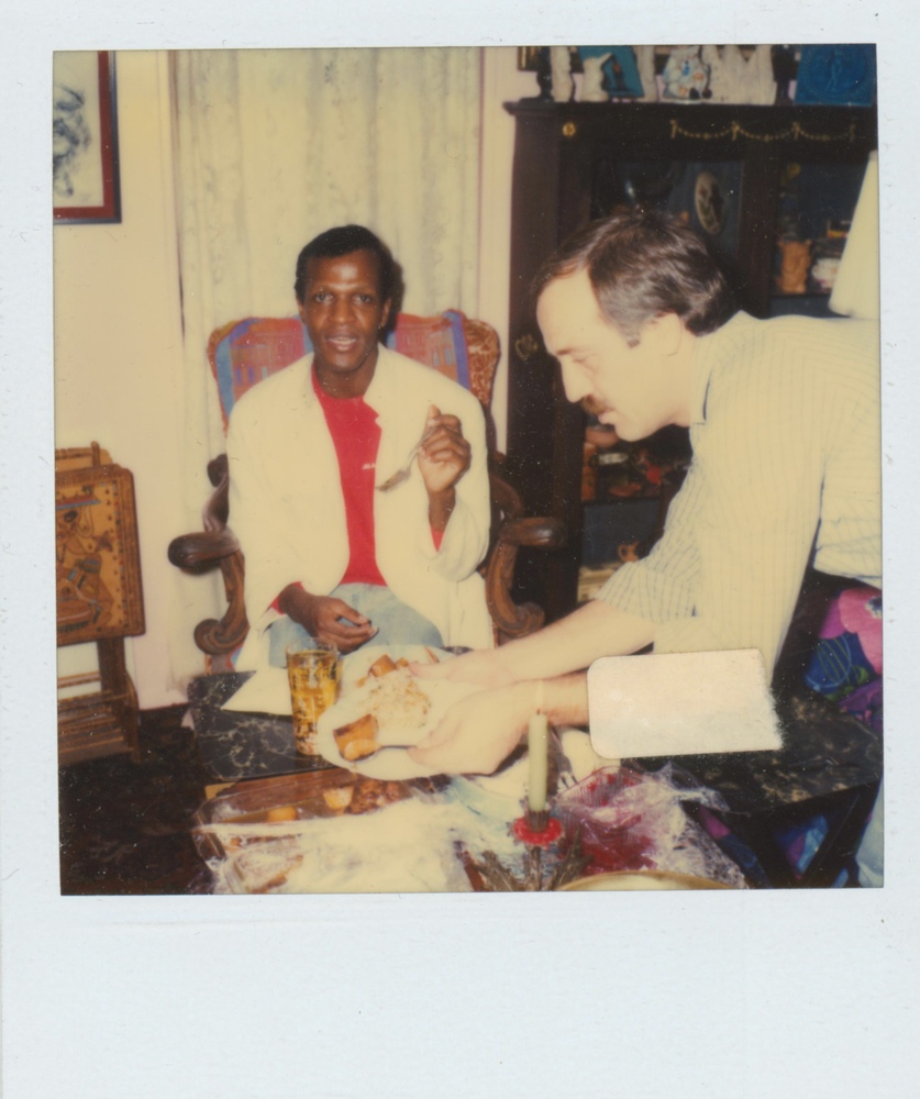 Download the full-sized image of A Photograph of Marsha P. Johnson Sitting with a Fork In Her Hand, Eating Food a Coffee Table