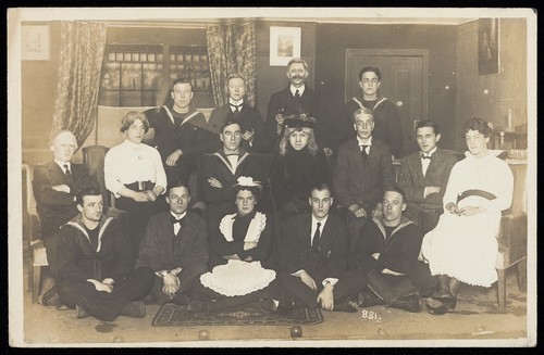 Download the full-sized image of Sailors, some in drag, posing for a group portrait within a highly-detailed stage set. Photographic postcard, 191-.